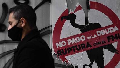 A man in a face mask walks in front of a sign that reads "No pago de la deuda Ruptura con el FMI"