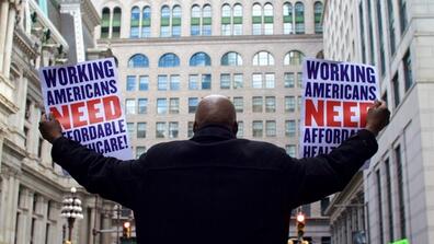 A man holds up signs reading "Working Americans Need Affordable Healthcare!"