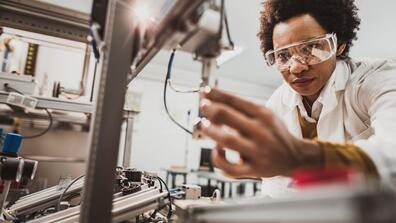 A scientist wearing lab goggles leans over to adjust a device.